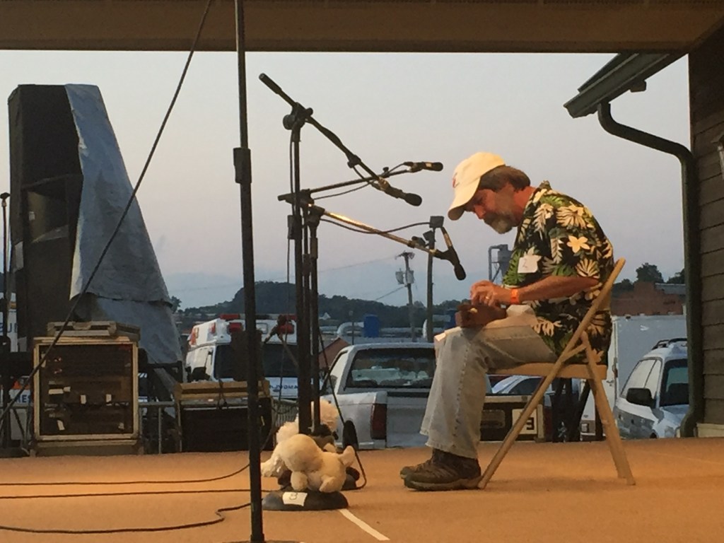 Man in a Hawaiian shirt, blue jeans and while ball cap playing a dulcimer.